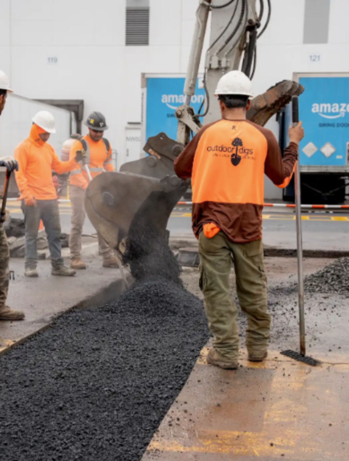Workers applying asphalt on a construction site with an Amazon truck in the background.